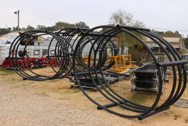 Large, cylindrical black metal frames are stacked outdoors on gravel. Background includes construction equipment and industrial buildings, under an overcast sky.