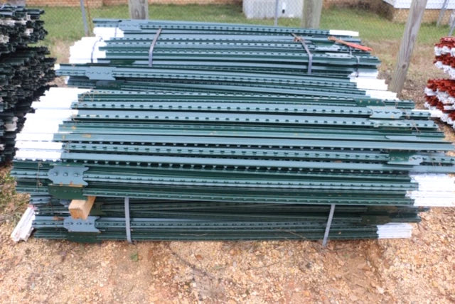 A stack of green metal fence posts, bound with straps, lies on a gravelly ground. Posts are neatly organized, conveying a sense of order and readiness for use.
